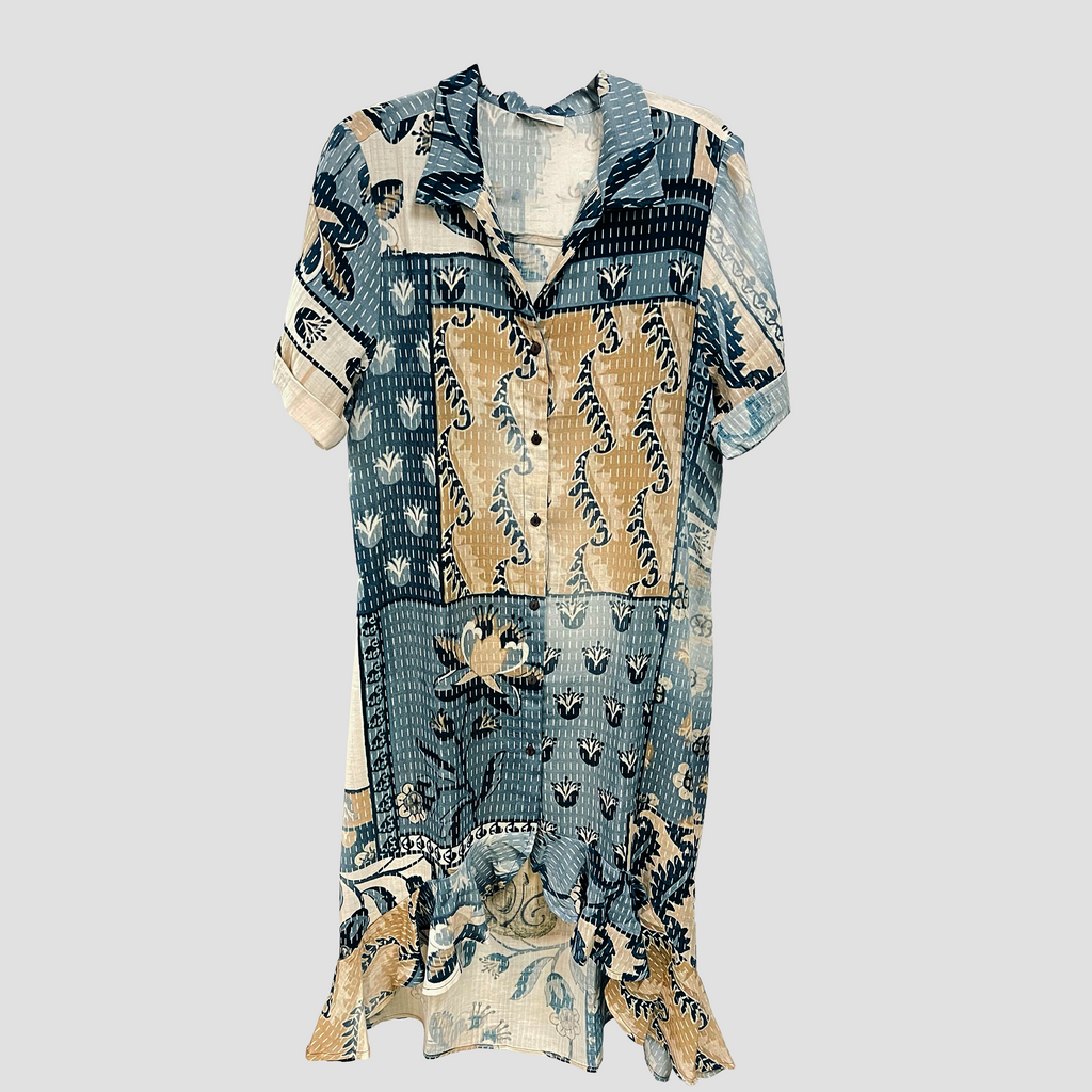 A short-sleeve button-up shirt dress with a patchwork batik print in shades of blue, beige, and white, displayed against a solid light gray background.