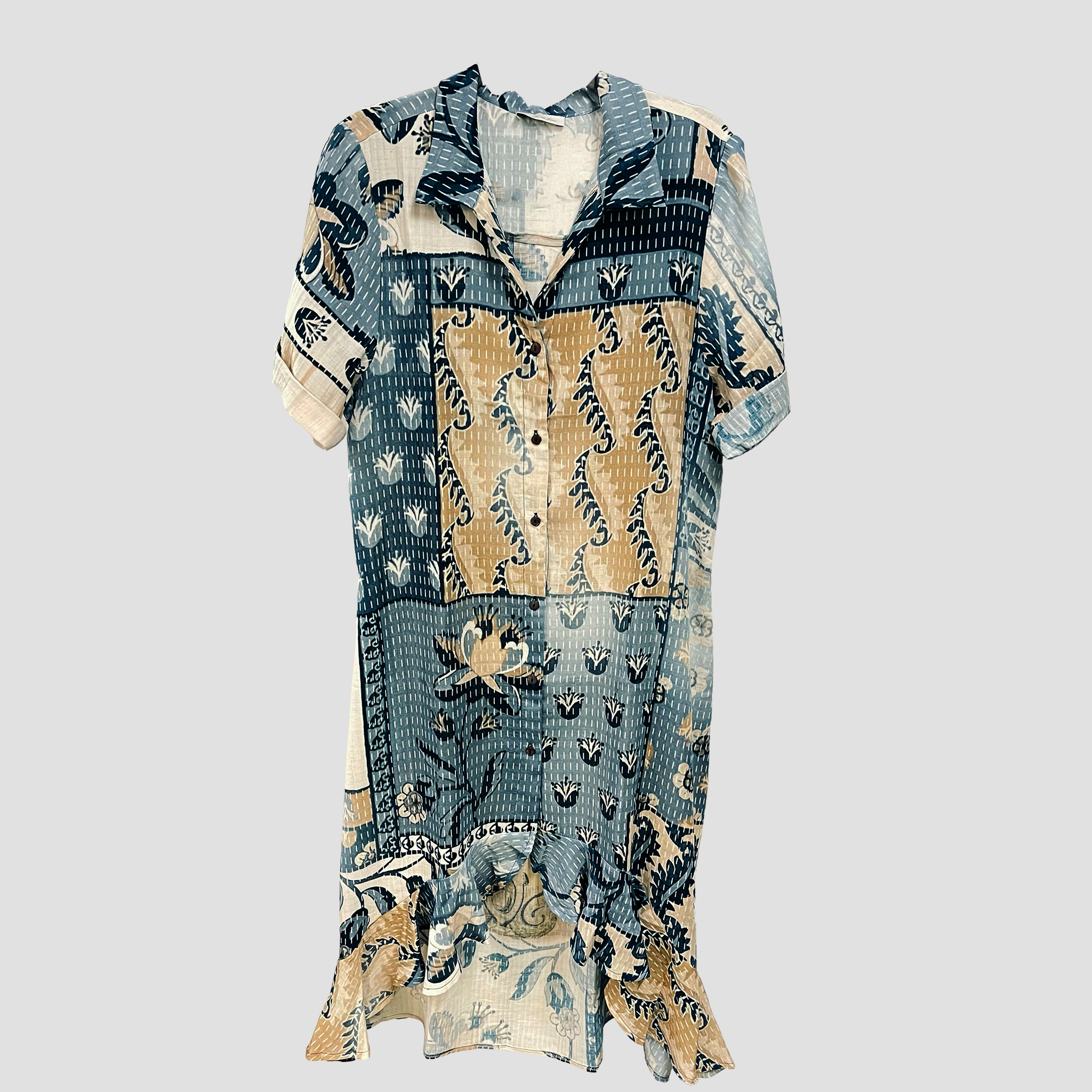 A short-sleeve button-up shirt dress with a patchwork batik print in shades of blue, beige, and white, displayed against a solid light gray background.