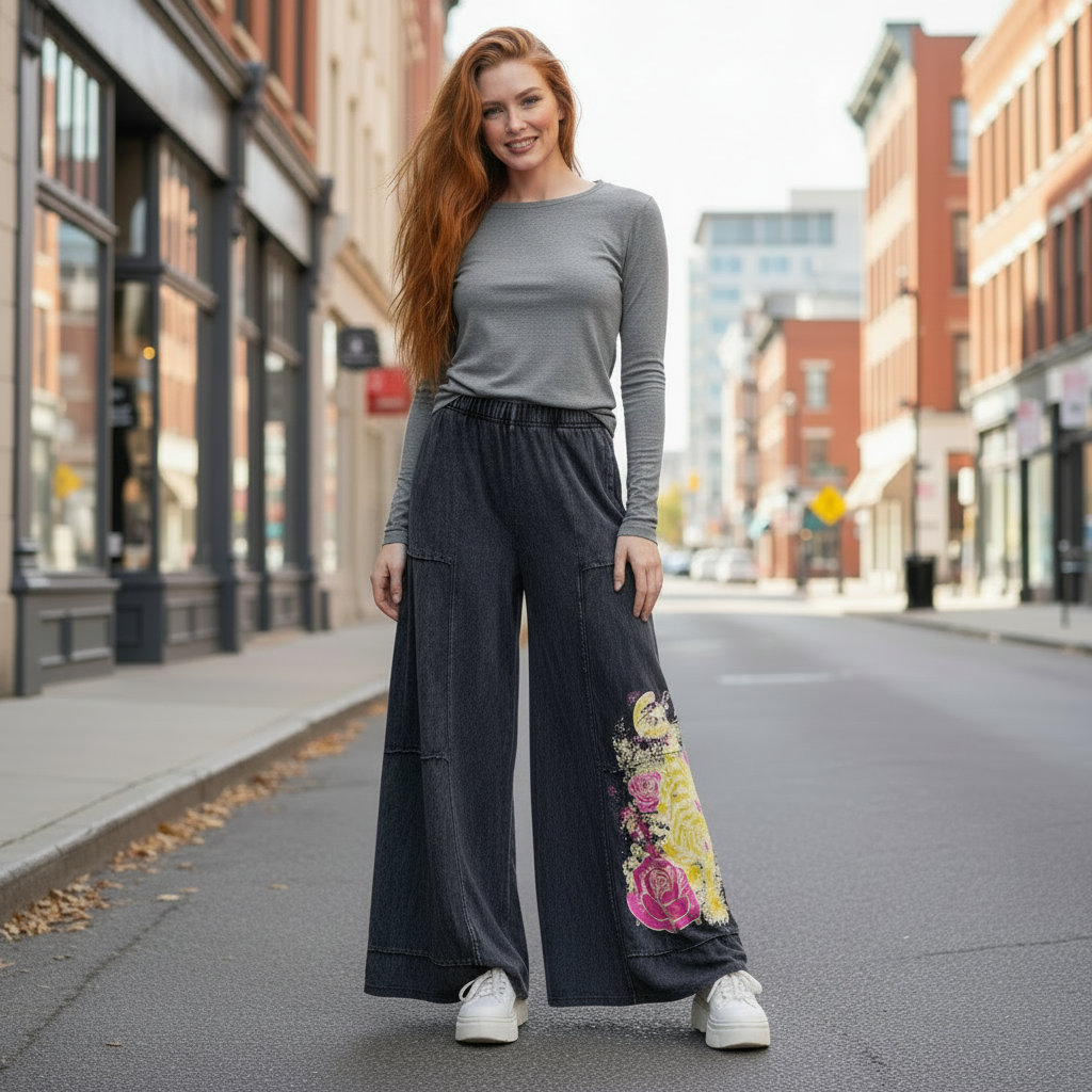 A woman stands on an urban street wearing a gray long sleeve top, wide-leg dark gray pants with a colorful floral print near the lower right leg, and white sneakers.